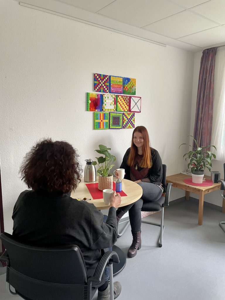 Zwei Personen sitzen an einem runden Tisch in einem gemütlichen Raum mit Topfpflanzen. Die eine hat lange rote Haare und lächelt mit einer Tasse in der Hand, die andere hat kurze, lockige Haare. An der Wand hinter ihnen hängt ein farbenfrohes Kunstwerk.