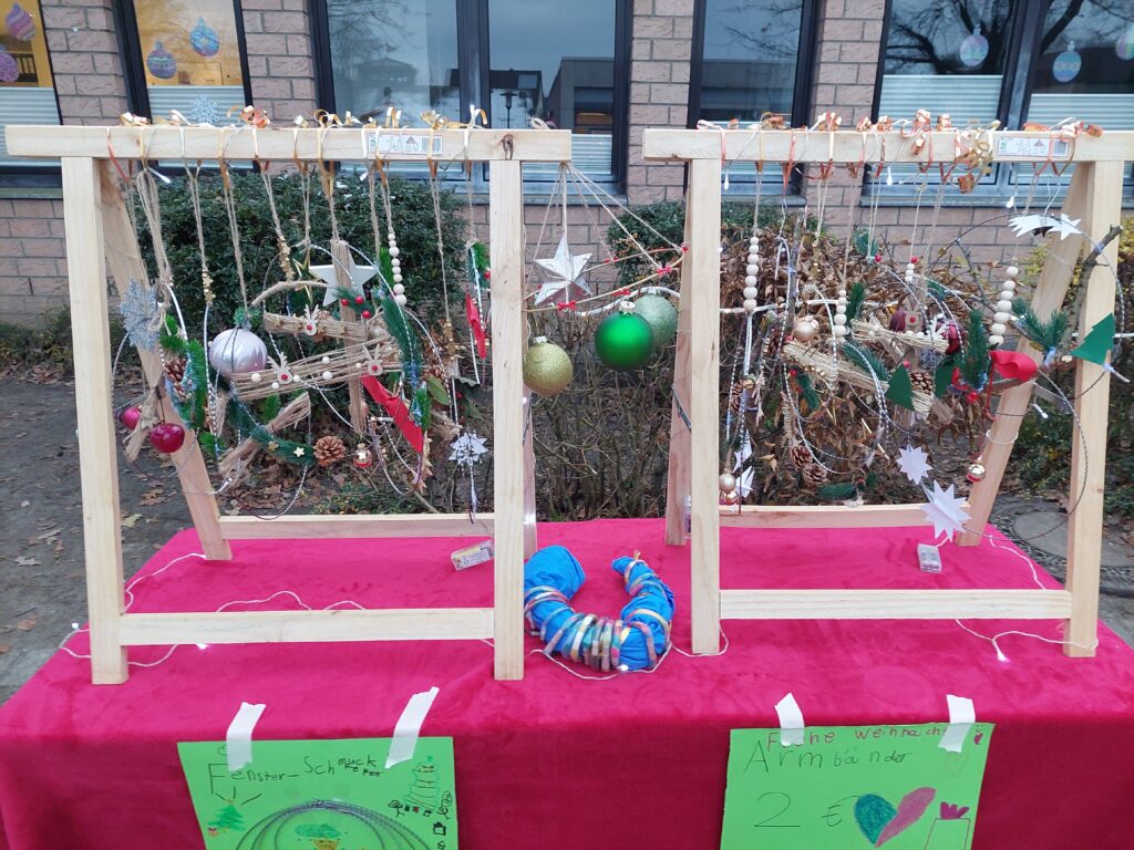 Festliche Präsentation mit zwei Holzrahmen, geschmückt mit bunten Ornamenten, Sternen und Grünpflanzen. Darunter liegt ein rotes Tischtuch, auf dem Kinderzeichnungen und ein Schild mit dem Hinweis auf Armbänder für 2 € zu sehen sind. Ein Armband liegt auf dem Tuch.