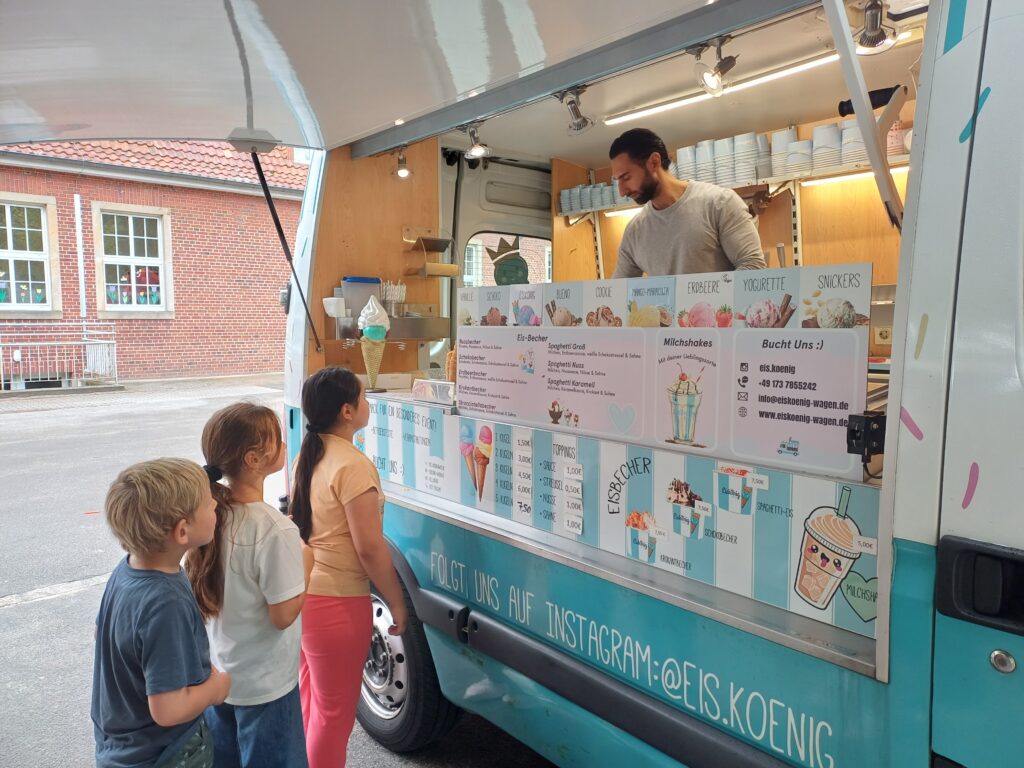 Drei Grundschulkinder stehen vor einem bunten Eiswagen Schlange und warten geduldig auf ihr Eis, welches gerade von einem Mitarbeiter des Eiswagens portioniert wird.