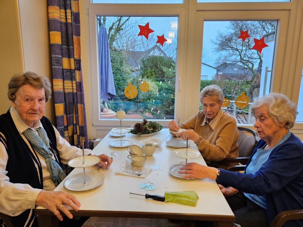 Drei Seniorinnen sitzen gemeinsam an einem Tisch in der Tagespflege Laggenbeck und basteln Etageren aus Geschirr. Auf dem Tisch liegen Teller, Tassen und Bastelmaterial, im Hintergrund sind Fenster mit winterlicher Dekoration zu sehen.