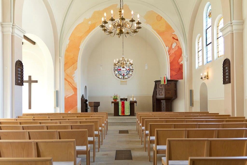 Ein ruhiger Kirchenraum mit Holzbänken vor einem mit bunten Tüchern geschmückten Altar. Buntglasfenster säumen die Wände, und ein großer Kronleuchter hängt von der Decke. Ein Kreuz schmückt die weiße Wand links.