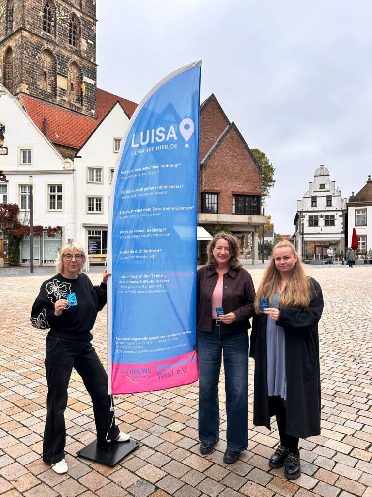 Drei Frauen stehen auf einem Platz in Rheine neben einer blauen Fahne mit der Aufschrift „Luisa ist hier“. Sie halten blaue Schutz-Deckel für Getränke in den Händen. Die Aktion informiert über Hilfe in bedrohlichen Situationen und den Schutz vor K.O.-Tropfen.