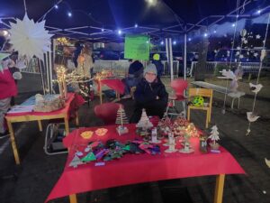 Ein festlicher Marktstand im Freien unter einem Baldachin, dekoriert mit Lichterketten. Verschiedene handgefertigte Gegenstände, darunter Dekorationen und Ornamente aus Holz, werden auf roten Tischdecken präsentiert. Eine Person sitzt lächelnd hinter dem Tisch.
