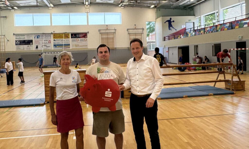 Stephan Runde von der Kreissparkasse Steinfurt überreicht in der Willibrordhalle einen symbolischen Spendenscheck über 1.500 Euro an Marius Wittenberg vom Offenen Ganztag und Dörte Michels vom TB Burgsteinfurt. Im Hintergrund sind Kinder beim Sportangebot in der Turnhalle zu sehen.