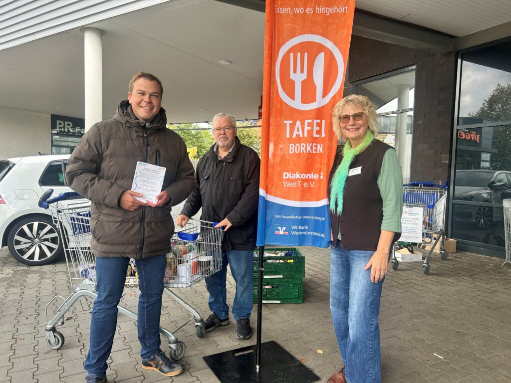 Drei Personen stehen beim Aktionstag der Tafel Borken vor einem Supermarkt. Neben ihnen sind eine Tafel-Fahne, Einkaufswagen mit gespendeten Lebensmitteln und grüne Transportkisten zu sehen.