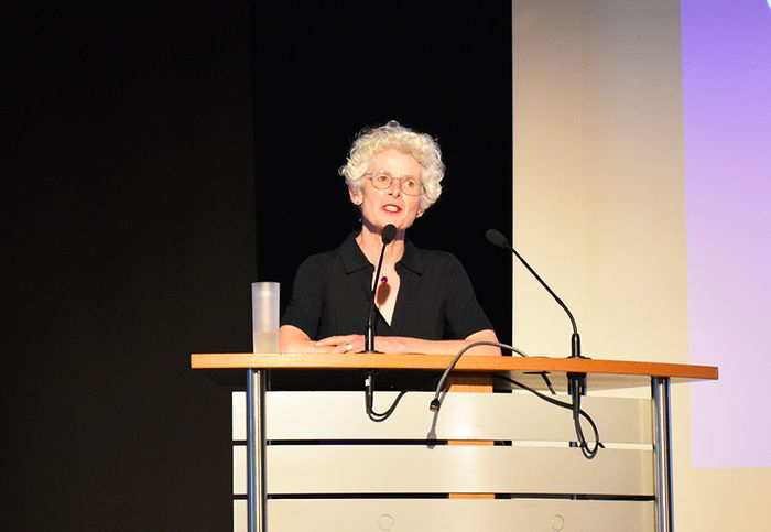 Imke Philipps steht an einem Rednerpult und spricht bei einer Synodenveranstaltung. Sie trägt dunkle Kleidung und spricht in ein Mikrofon. Im Hintergrund ist eine Leinwand zu sehen.